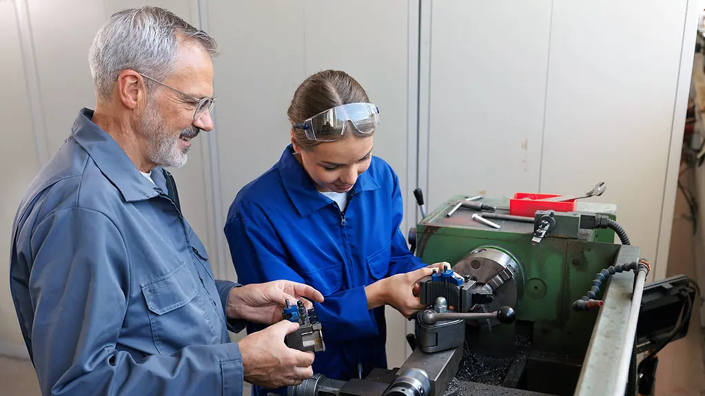 Auszubildende bei der Arbeit mit einem Ausbilder, der neben einer Drehmaschine in einer Werkstatt steht.