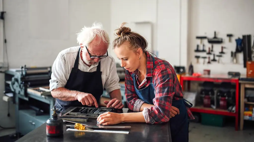 An older male colleague and a young female colleague are standing together at a desk.