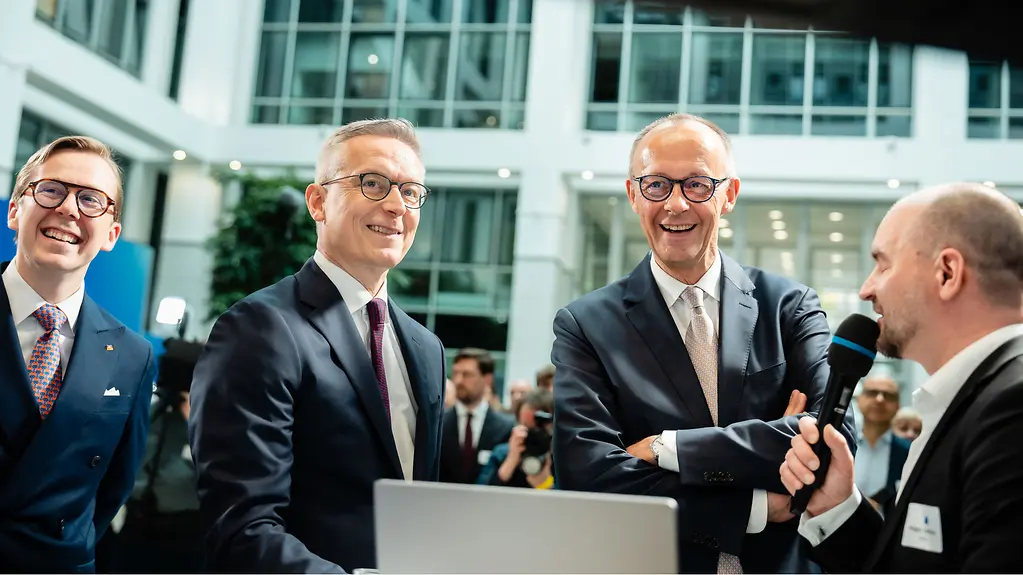 Bundeskanzler Merz und Bundesminister Wildberger lachen bei einer Veranstaltung zum einjährigen Bestehen des Ministeriums.