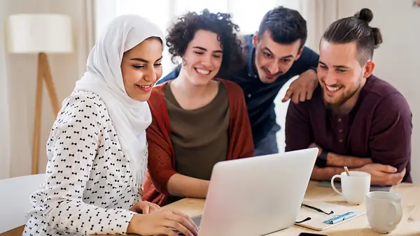 Eine multiethnische Gruppe junger Freunde sitzen und stehen vor einem Laptop.