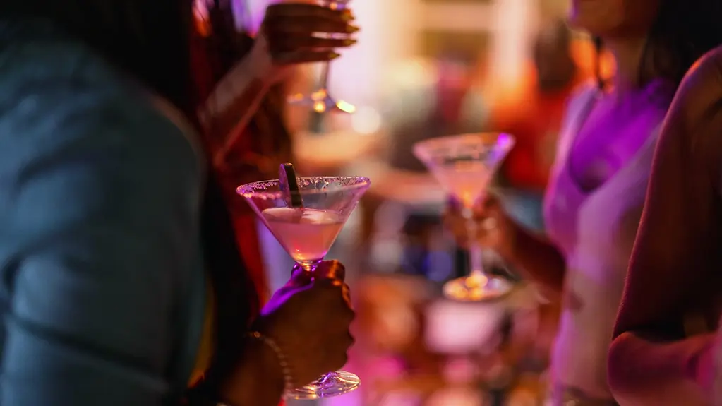 Nahaufnahme eines Martiniglases, das von einer Frau an einer Bar mit violetter Beleuchtung gehalten wird.