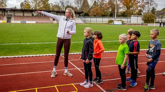 Auf einem Sportplatz gibt eine sportlich gekleidete Person Anweisungen an eine kleine Gruppe ebenfalls sportlich bekleidete Kinder