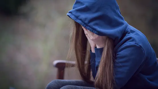 Ein Mädchen sitzt auf einer Bank und hält die Hände vor das Gesicht. Sie hat die Kapuze eines Hoodies auf dem Kopf. Es schauen nur ihre langen Haare heraus.