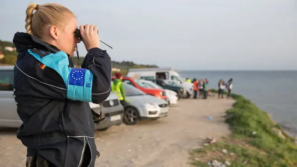Eine deutsche Bundespolizistin mit Frontex-Armbinde steht an der Küste und schaut durch ein Fernglas aufs Meer.