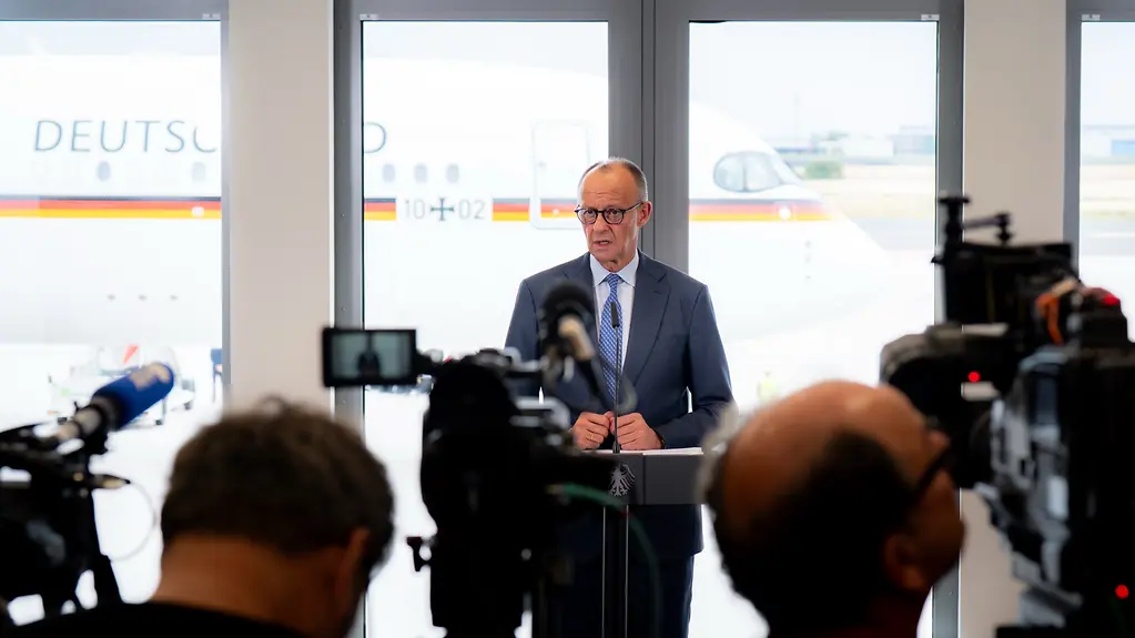 Kanzler Merz spricht am Flughafen, im Hintergrund steht der Regierungsflieger.