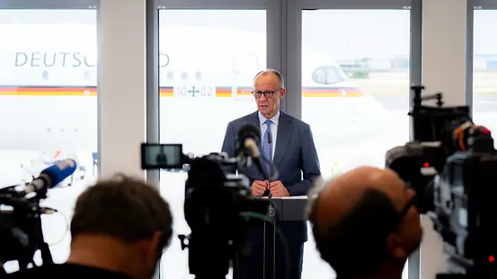 Kanzler Merz spricht am Flughafen, im Hintergrund steht der Regierungsflieger.