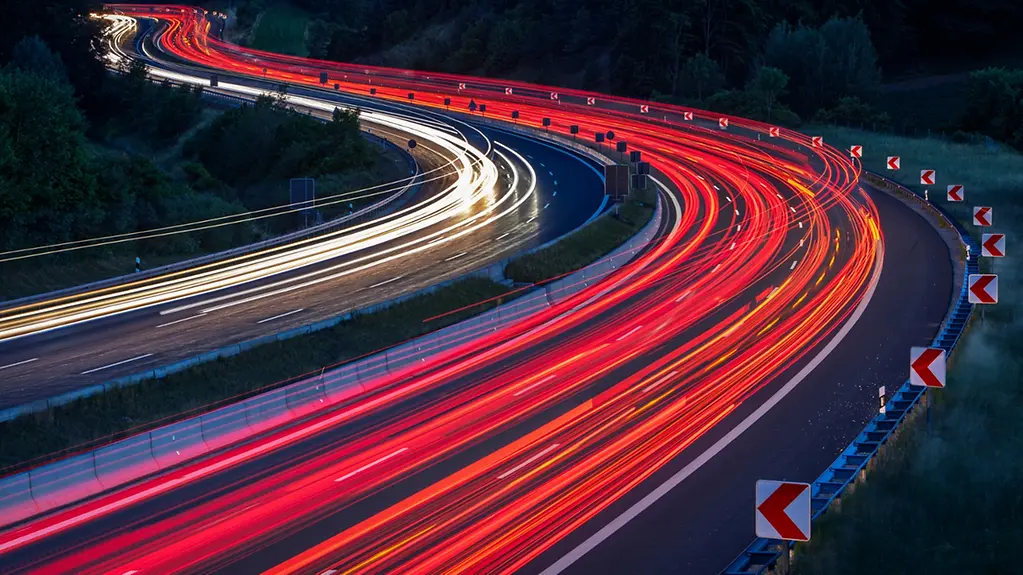 Abstrakte rote und weiße Lichtspuren von PKWs und LKWs auf einer mehrspurigen, kurvenreichen Autobahn, inmitten grüner Wälder und Felder bei Nacht