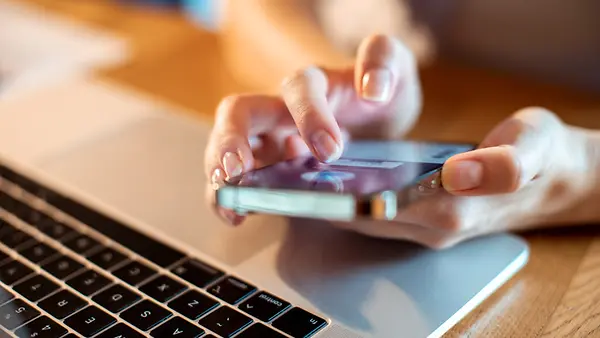 Eine Frau hat ein Smartphone in der Hand, ein aufgeklappter Laptop ist ebenfalls zu sehen. 