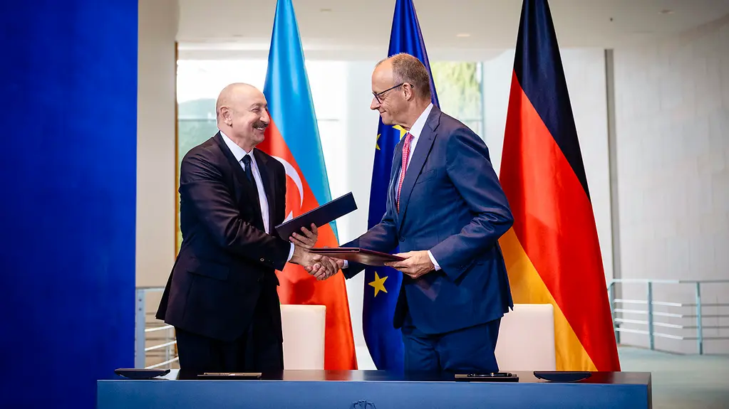 Federal Chancellor Merz and President Aliyev shaking hands, holding signed documents in their hands.