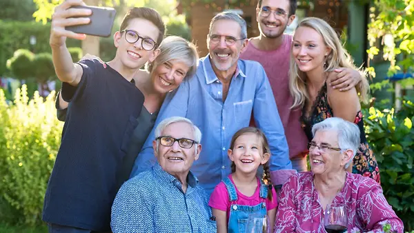 Im Sommer hat eine Familie von drei Generationen Spaß an einem Tisch im Garten. Ein Junge nimmt ein Selfie von der Mehrgenerationen-Familie auf.