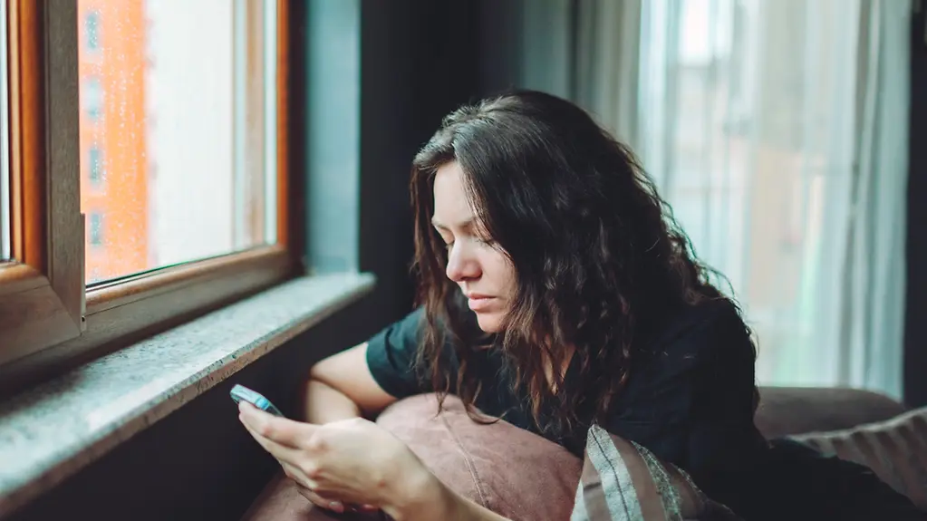 A woman is sitting on a sofa, looking exhausted as she stares at her mobile phone.