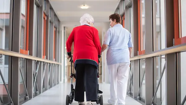 Eine Pflegerin geht mit einer Bewohnerin des Pflegeheims und einem Rollator über den Flur. 