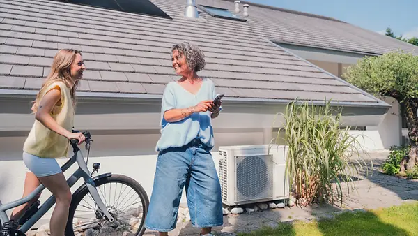 Mutter und Tochter mit Fahrrad unterhalten sich in der Nähe eines Hauses mit Solaranlage und Wärmepumpe.