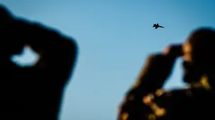 Am Himmel fliegt ein Kampfflugzeug. Im Vordergrund stehen schemenhaft zwei Personen mit Fernglas, die das Flugzeug beobachten.