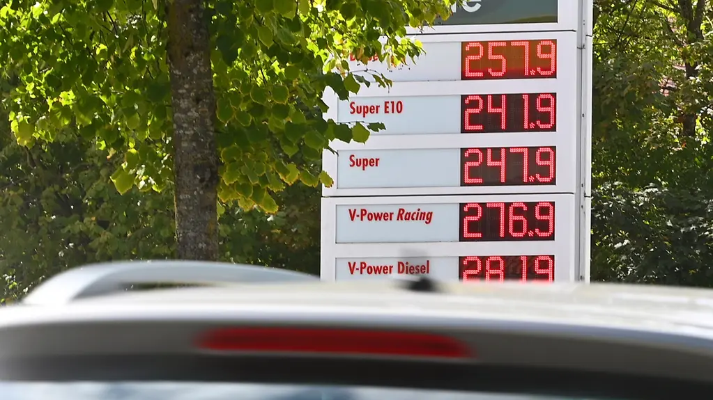 Hinter einem Auto steht die Anzeigetafel einer Tankstelle, auf der man die aktuellen Spritpreise sieht.