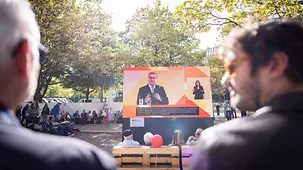Bürgerinnen und Bürger vor einem Livestream das einen Talk zeigt.