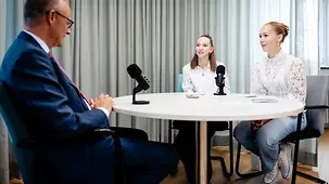 Bundeskanzler Friedrich Merz mit Schülerinnen an einem Tisch, vor ihnen Mikrofone.