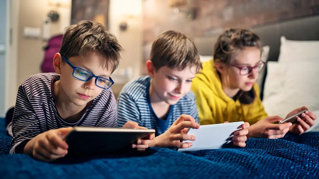 Three children are lying on a bed, looking at their mobile phones and tablets.