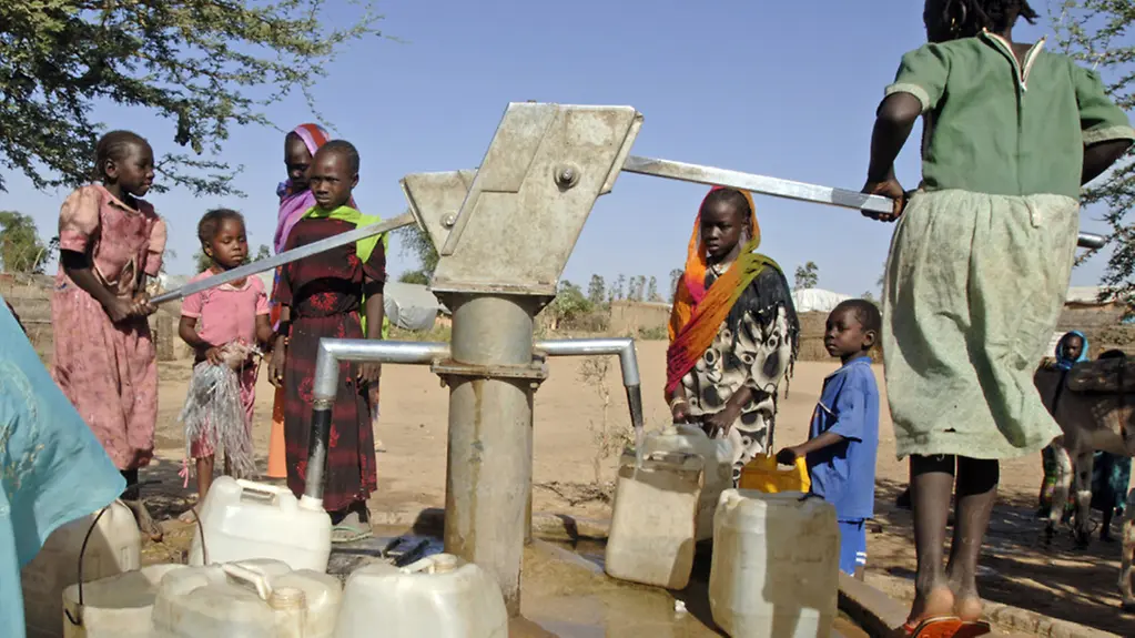 Kinder und Frauen an einer Hand-Wasserpumpe.
