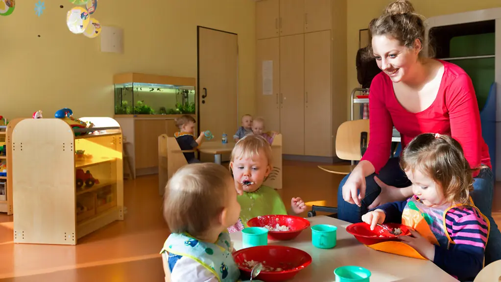 Drei Kita-Kinder sitzen an einem kleinen Tisch und Essen. Eine Erzieherin betreut sie.