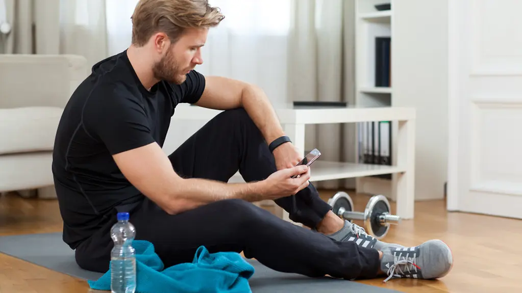 Ein junger Mann vergleicht die Daten seines Fitnessarmbands mit denen seiner Sport-App auf dem Smartphone.
