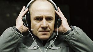 Ulrich Mühe mit Kopfhörern auf dem Ohren beim Überwachen einer Künstlerfamilie