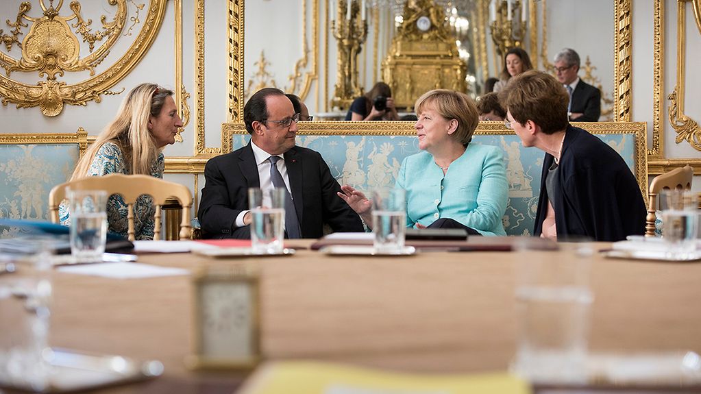 Assis sur un divan, Angela Merkel et François Hollande sont en train de discuter ; à leurs côtés (à d. et à g.) leurs interprètes