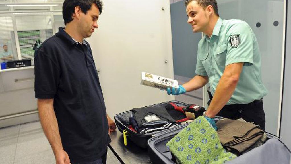 Ein Zollbeamter hat den Koffer eines Fluggastes durchsucht und zeigt ihm eine im Koffer gefundene Stange mit Zigaretten.