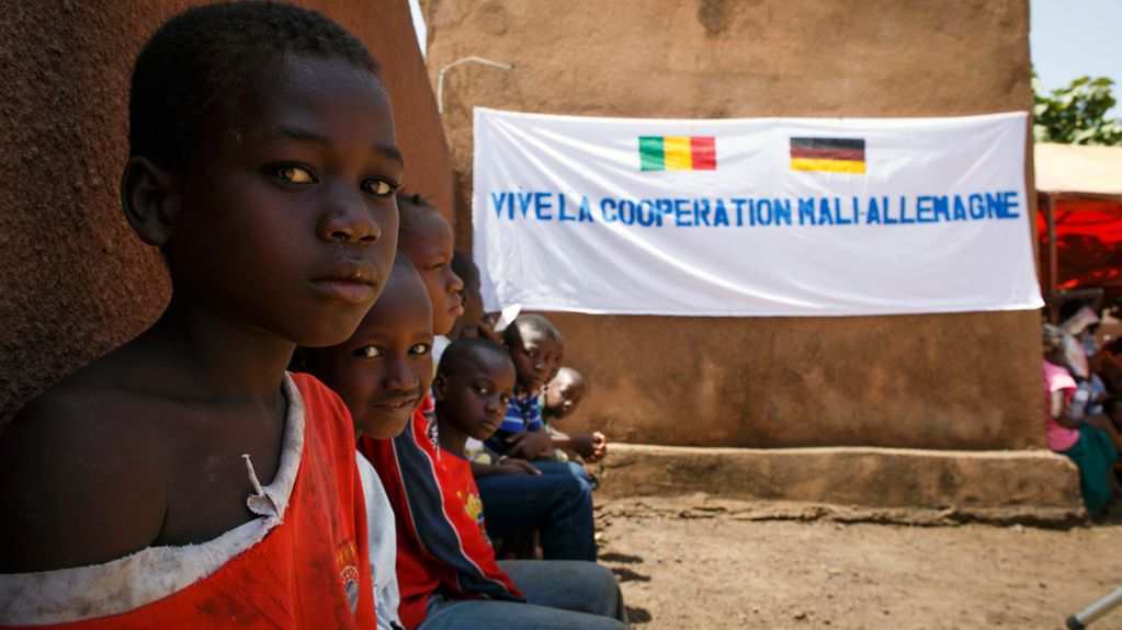 Kinder sitzen in der Gemeinde Tienfala vor einem Banner mit der Aufschrift ' Es lebe die Deutsch-Malische Kooperation'. Tienfala, Mali. 28.03.2014. Copyright: Thomas Trutschel/ photothek.net