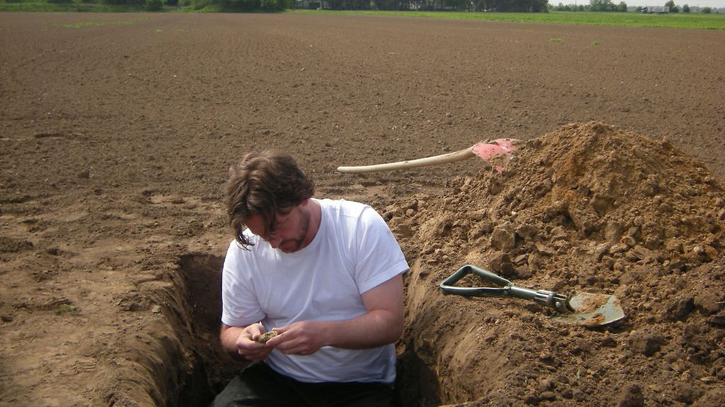 Ein BGR-Bodenkundler katalogisiert im Feld den zu beprobenden Sandboden. Die Probe wird aus dem Oberboden entnommen, der sich durch seinen Humusgehalt deutlich vom ockerfarbenen Unterboden unterscheidet.