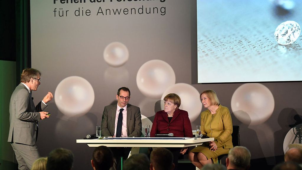 Kanzlerin Merkel und Ministerin Wanka sitzen an einem Tisch und schauen Herrn Ambacher an.