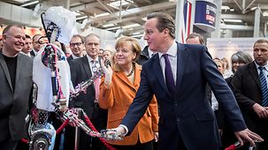 Bundeskanzlerin Angela Merkel und der britische Premierminister David Cameron an einem Stand mit einem Roboter.
