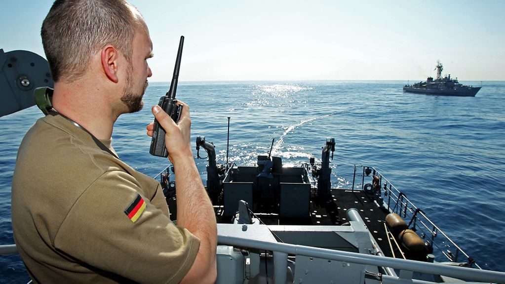 Soldat an Bord eines Schiff, spricht in Funkgerät, im Hintergrund ein Schiff