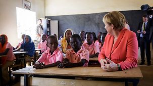 Bundeskanzlerin Angela Merkel sitzt beim Besuch einer Schulklasse an einer Schülerbank neben Kindern.