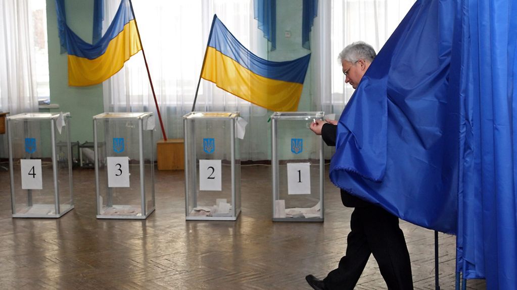 Mann verlässt Wahlkabine mit blauem Vorhang in einem Wahllokal mit Urnen und ukrainischen Fahnen.
