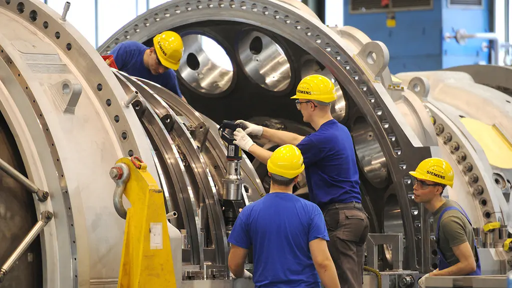 Vier Techniker arbeiten in einer Lagerhalle an der leistungsstärksten Gasturbine der Welt