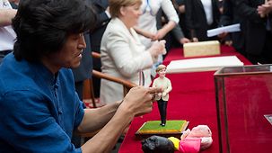 Ein Künstler fertigt eine kleine Tonfigur von Bundeskanzlerin Angela Merkel, die im Hintergrund sitzt.