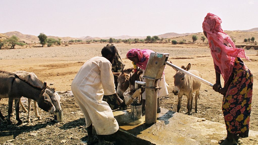 Menschen und Esel in Wüstenlandschaft an einer Wasserpumpe.