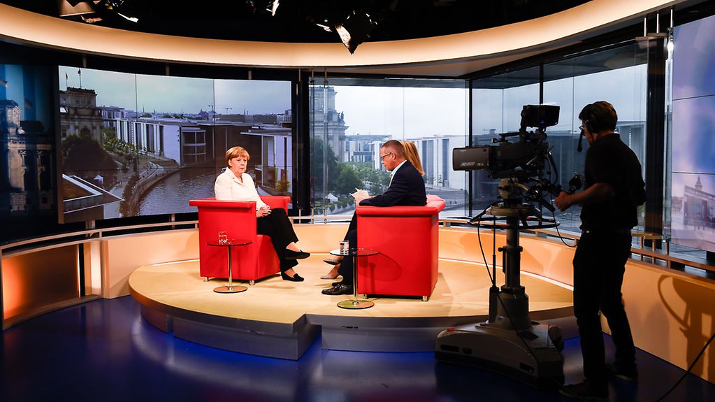 Fernsehstudio in der Totalen: links sitzend Kanzlerin Merkel, vorn rechts Kameramann