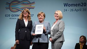 Bundeskanzlerin Angela Merkel mit dem Abschluss-Kommuniqué des Woman20 Summits zwischen Mona Küppers, Vorsitzende des Deutschen Frauenrates, und Stephanie Bschorr, Präsidentin des Verbandes Deutscher Unternehmerinnen e.V.