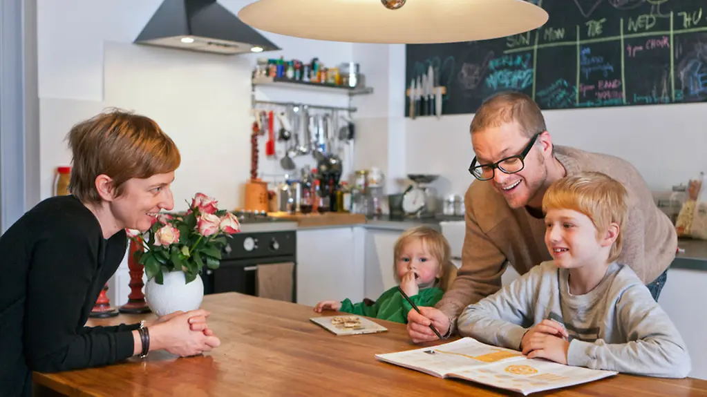 Zwei Kinder mit Eltern am Küchentisch, Vater hilft bei Schularbeiten.