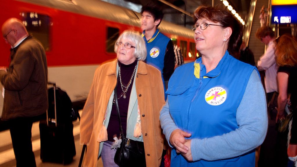 Ehrenamtliche Mitarbeiterin der Bahnhofsmission begleitet ältere Dame zum Zug.