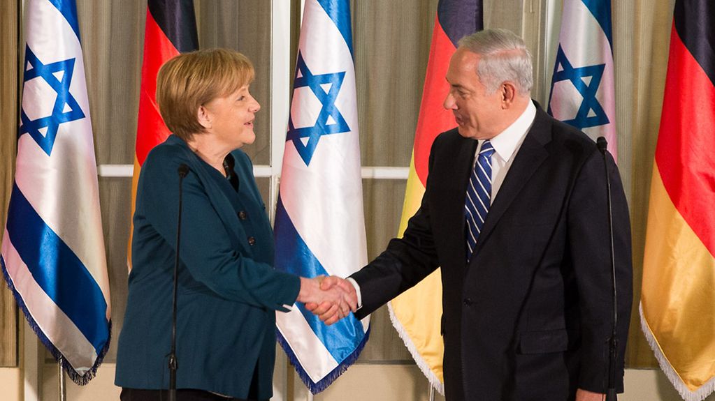 Chancellor Angela Merkel and Prime Minister Benjamin Netanjahu greet one another in front of German and Israeli flags.