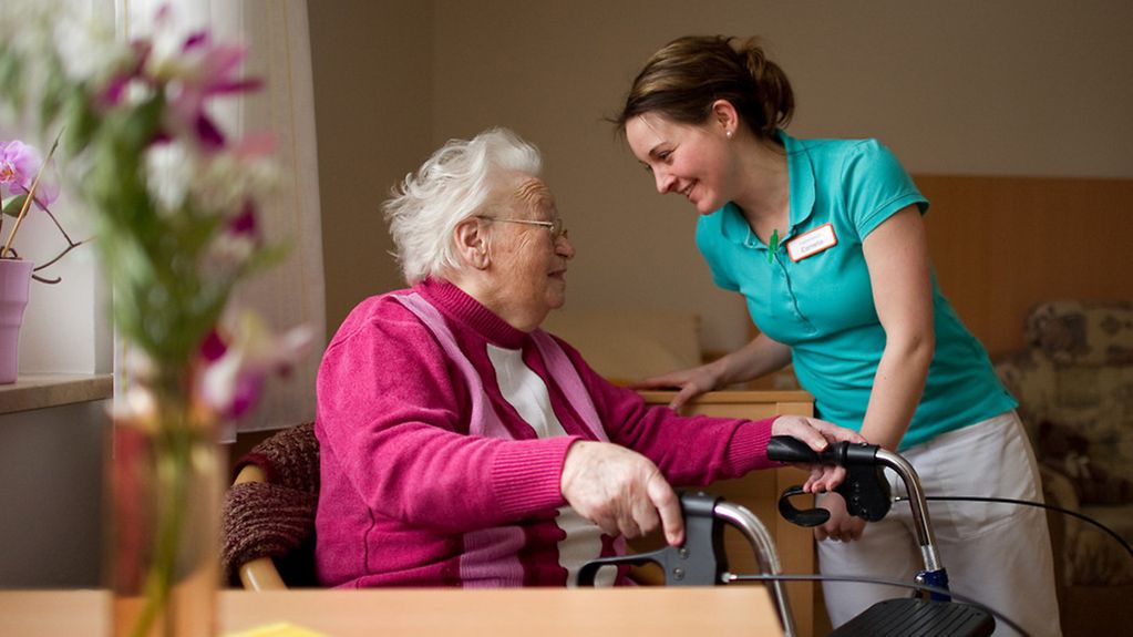 Pflegekraft unterhält sich mit älterer Dame im Rollstuhl im Zimmer.