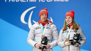 Die Athletin Clara Klug und ihr Guide Martin Härtl (beide Deutschland) stehen während der Siegerehrung auf der Bühne. Klug holte die Bronzemedaille im 10km Biathlon Damen sehbehindert.