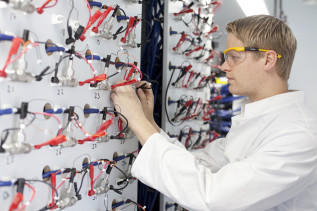 Ein junger Mann mit Laborkittel und Schutzbrille arbeitet an einer elektrischen Anlage. 