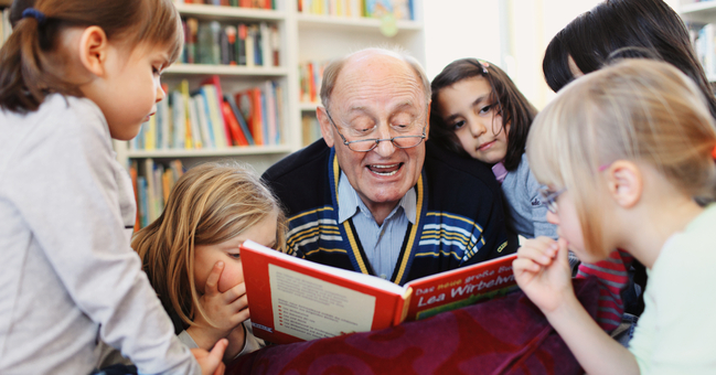 Ein älterer Mann liest aus einem Buch vor. Um ihn herum stehen fünf junge Mädchen.