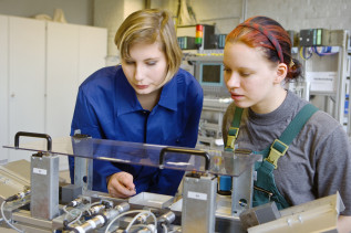 Zwei junge Frauen in Arbeitskleidung betrachten eine technische Anlage. 