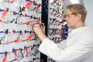Ein junger Mann mit Laborkittel und Schutzbrille arbeitet an einer elektrischen Anlage